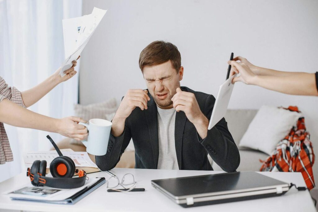 A young professional buried under papers, phone, and a laptop, representing overwhelm from saying yes to everything