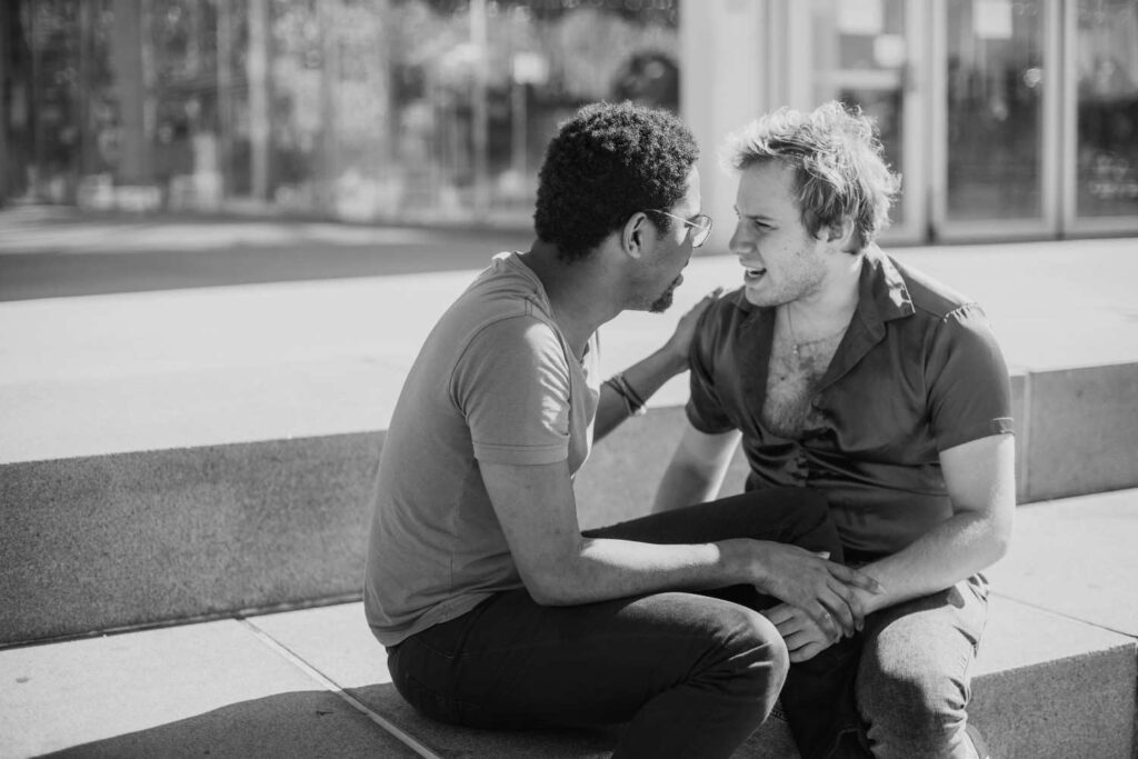 Two middle-aged men sitting on a park bench, one with hand on the other's shoulder in a supportive gesture, representing male friendship and emotional support

