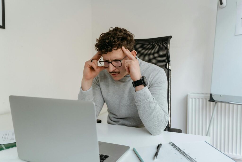 A young adult sitting at computer desk with hand on forehead, looking confused, representing brain fog symptoms, dehydration, and brain fog 2026
