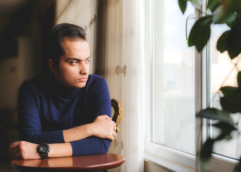 A young man sitting alone on floor looking out a rainy window, representing isolation and emotional struggle in toxic relationships
