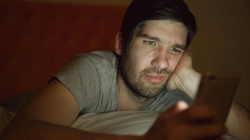 A young adult lying in bed scrolling on a smartphone at night with a concerned expression, blue light illuminating the face. phone addiction 2026
